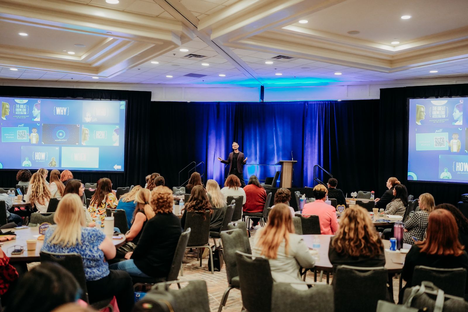 Luke Goetting speaking to a packed conference audience with dual screens